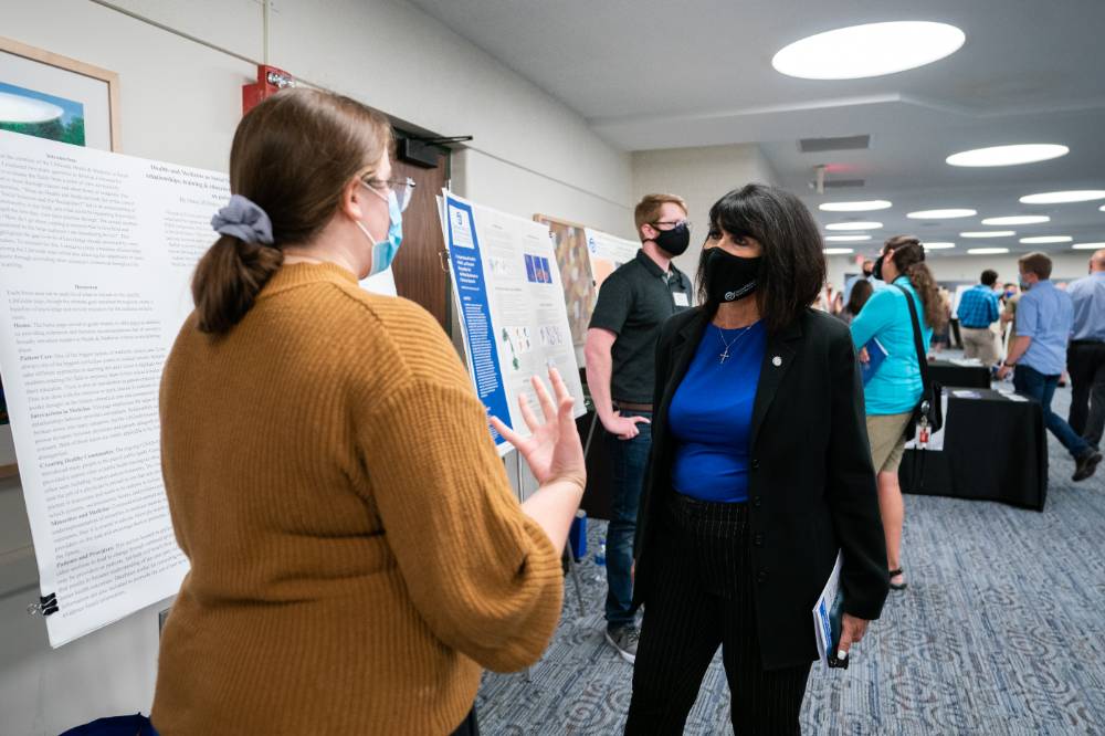 Scholar presenting poster to guest, both wearing masks.